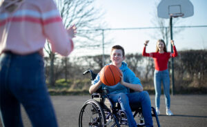 Teenage Boy In Wheelchair Playing Basketball With Friends